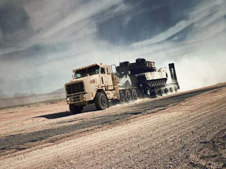 Oshkosh Defense Heavy Equipment Transporter (HET) driving on a sandy road with a military vehicle on the trailer.